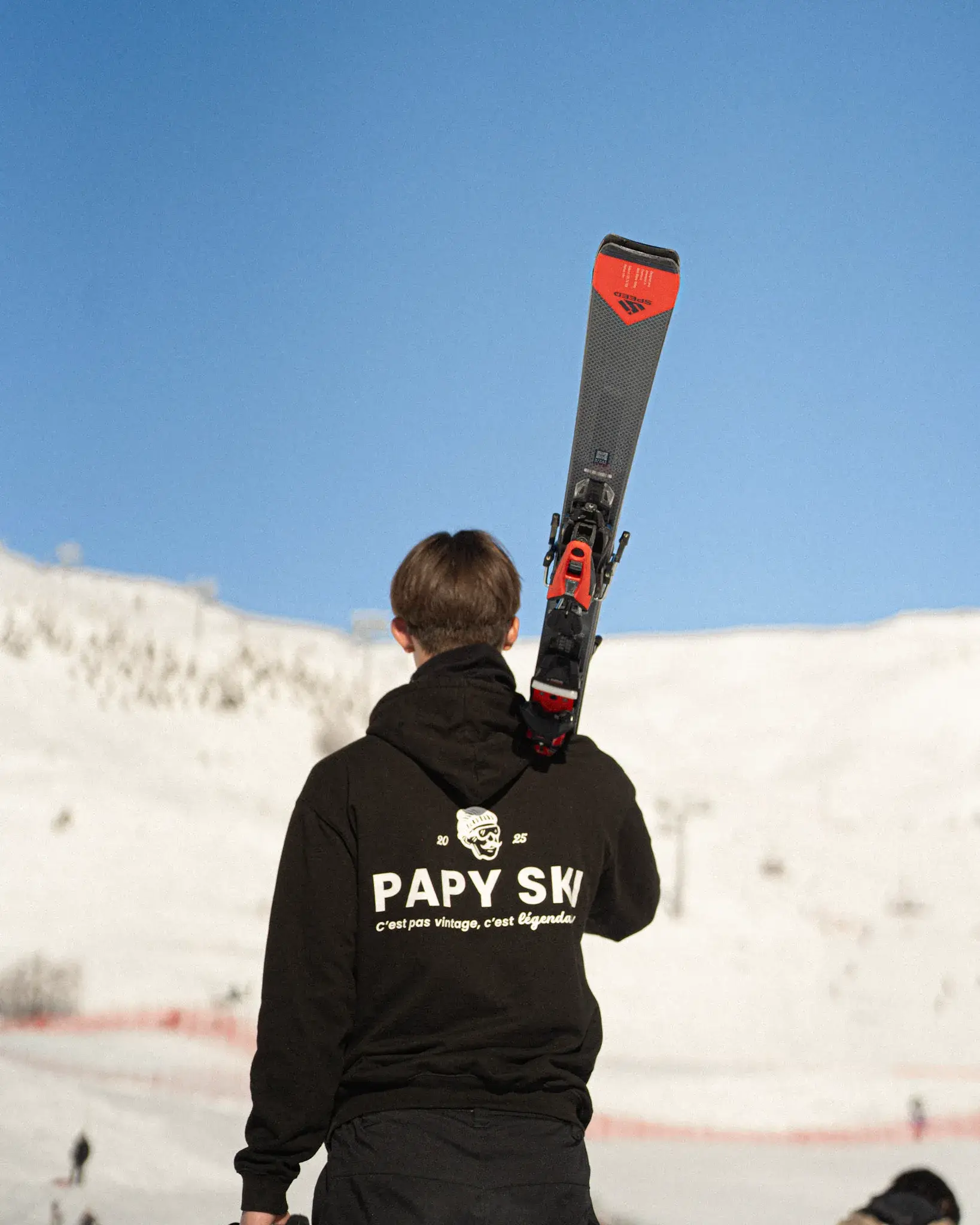 Skieur en sweat Papy Ski, skis sur l’épaule, face aux pistes d’Orcières